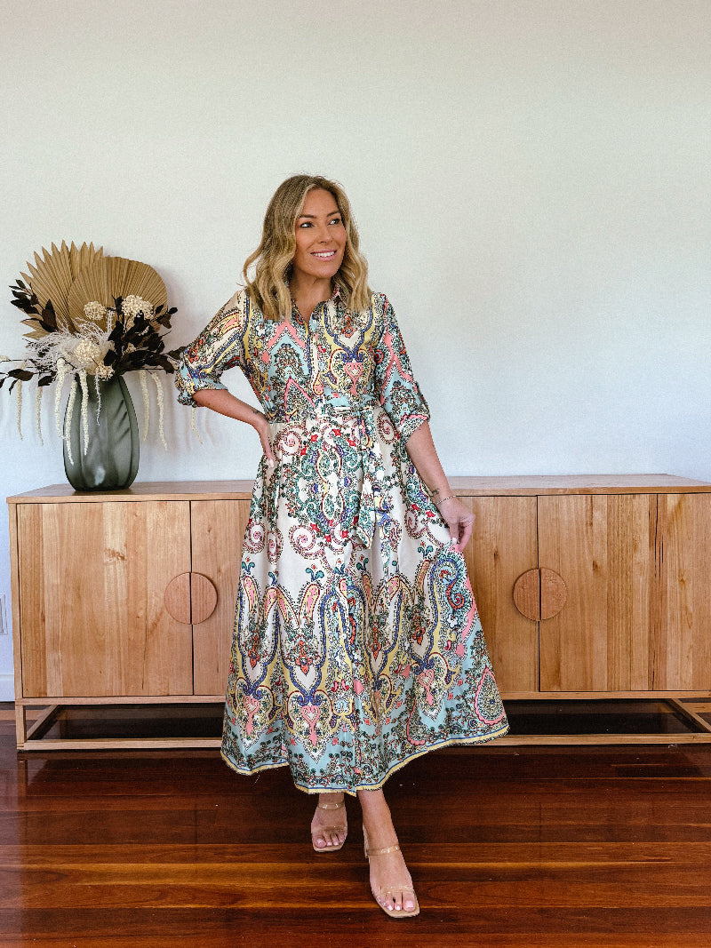 A woman standing in front of a wooden cabinet, wearing a long paisley shirt dress with 3/4 sleeves and flat sandals.
