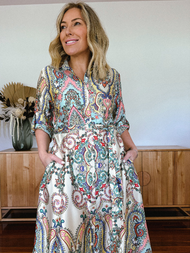 A woman standing in front of a wooden cabinet, wearing a long paisley shirt dress with 3/4 sleeves and flat sandals.